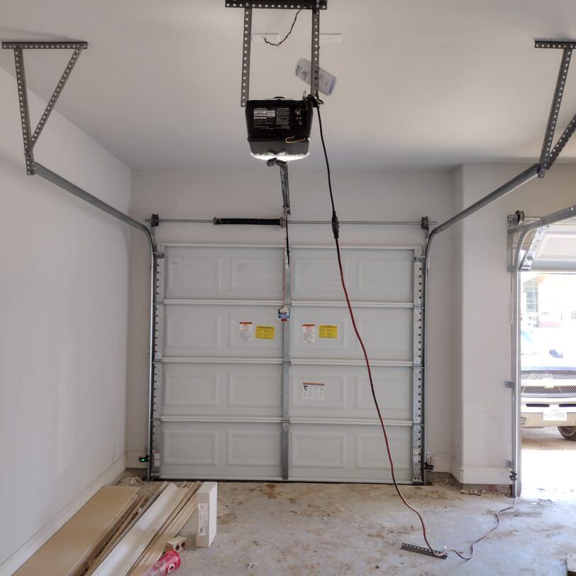 Garage interior with an open garage door, a garage door opener installed, and some building materials on the floor.