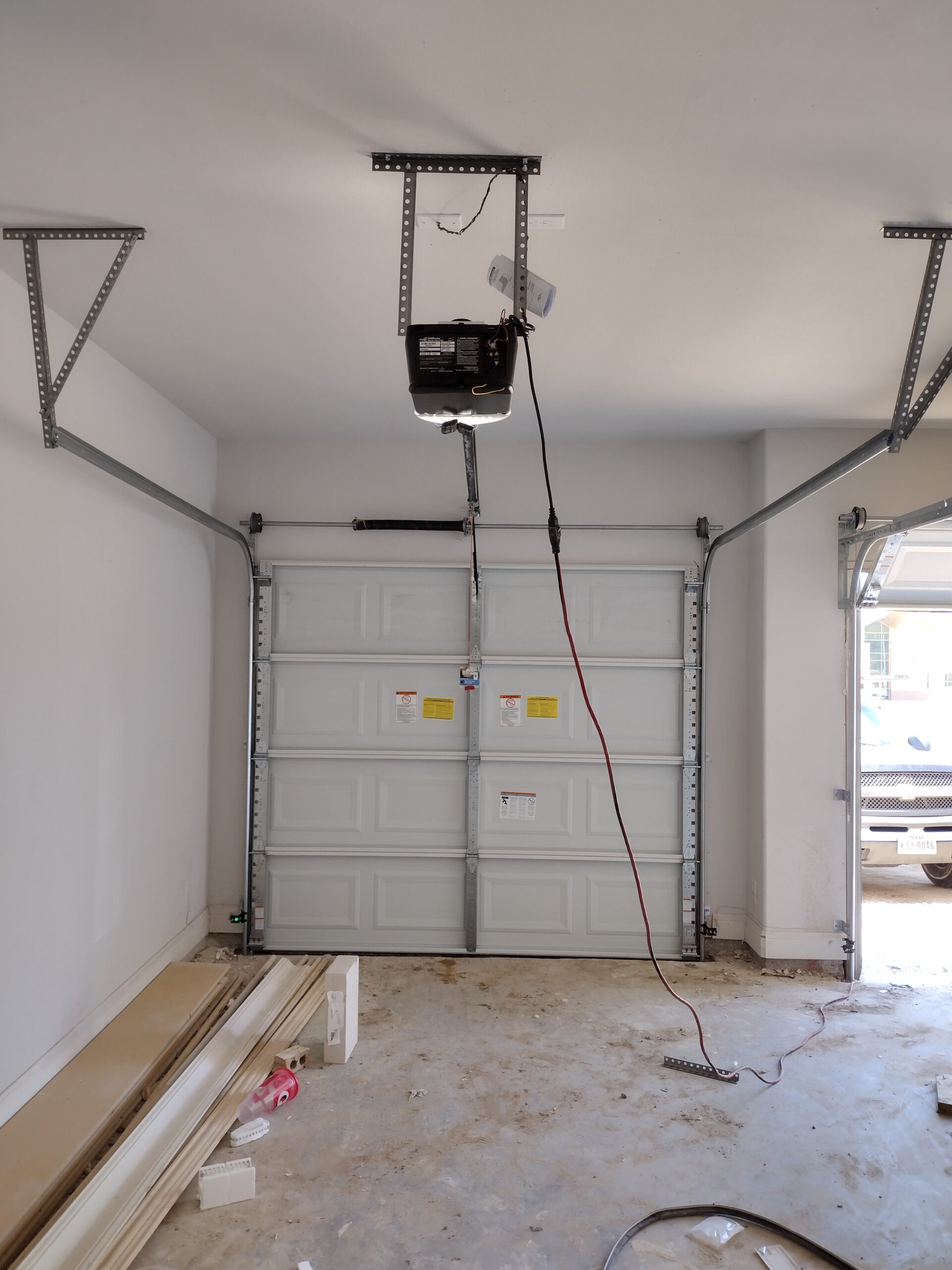 Garage interior with an open garage door, a garage door opener installed, and some building materials on the floor.