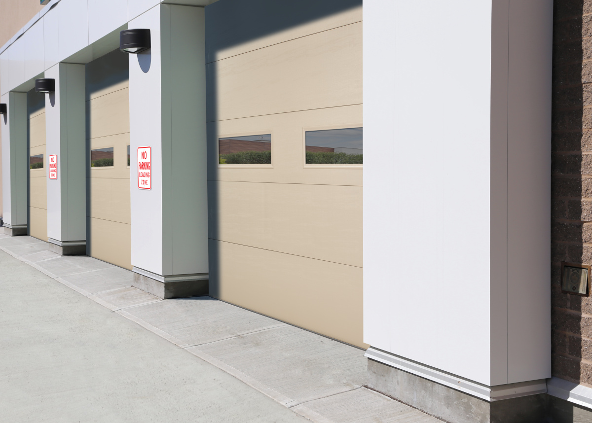 Modern commercial building exterior featuring multiple Amarr 2731 garage doors in a subtle beige color, each with small, rectangular windows offering a view of the countryside.