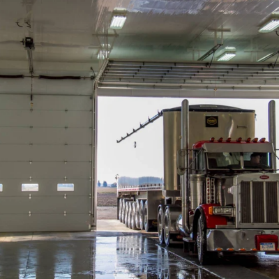 A large white commercial garage door open with a semi-truck pulling a grain trailer entering the building