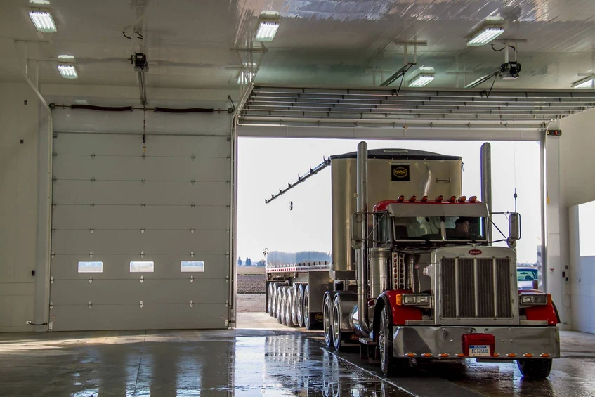 A large white commercial garage door open with a semi-truck pulling a grain trailer entering the building