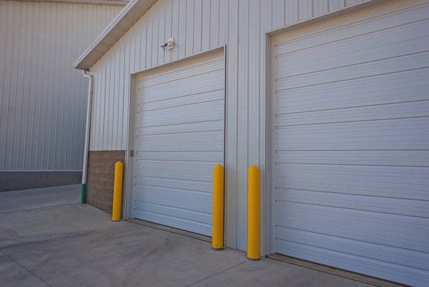 Two white commercial garage doors with a ribbed design, flanked by bright yellow protective bollards.