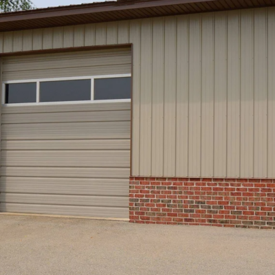 A beige commercial garage door with horizontal grooves and a row of windows at the top. The building has a brown roof and brick siding at the bottom.