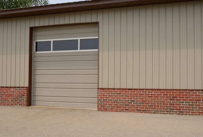 A beige commercial garage door with horizontal grooves and a row of windows at the top. The building has a brown roof and brick siding at the bottom.