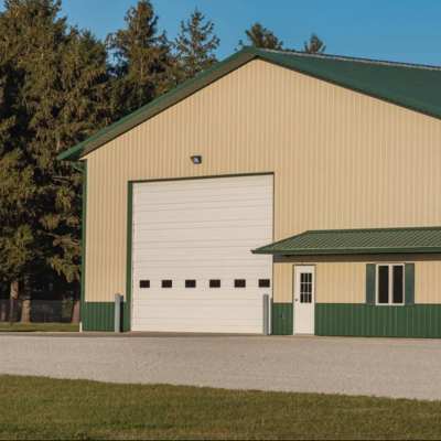 A large white commercial garage door on a beige metal building with a green roof and trim. The building is surrounded by green lawn and trees.