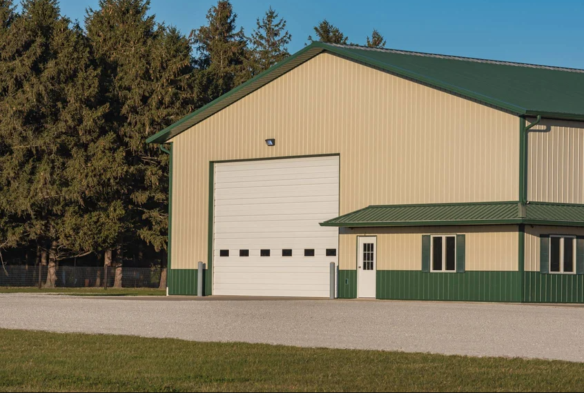 A large white commercial garage door on a beige metal building with a green roof and trim. The building is surrounded by green lawn and trees.