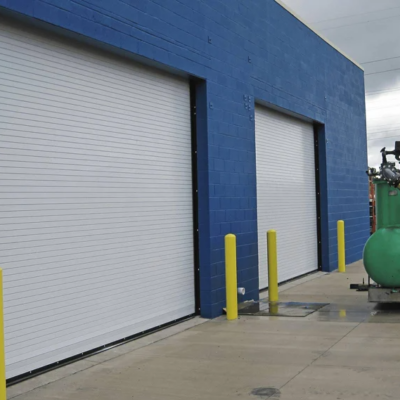 Two large white commercial roll-up doors on a blue building with yellow bollards in front.