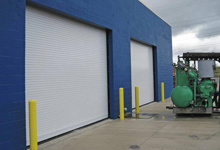 Two large white commercial roll-up doors on a blue building with yellow bollards in front.