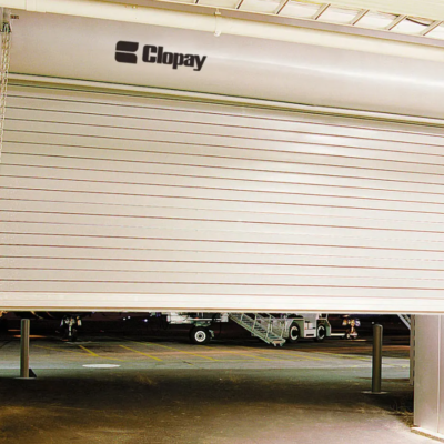 A partially opened Clopay rolling steel door revealing an airport tarmac with parked airplanes in the background.