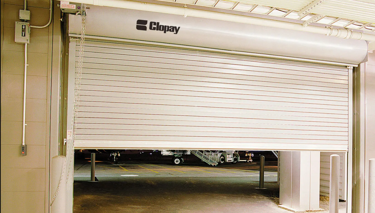 A partially opened Clopay rolling steel door revealing an airport tarmac with parked airplanes in the background.