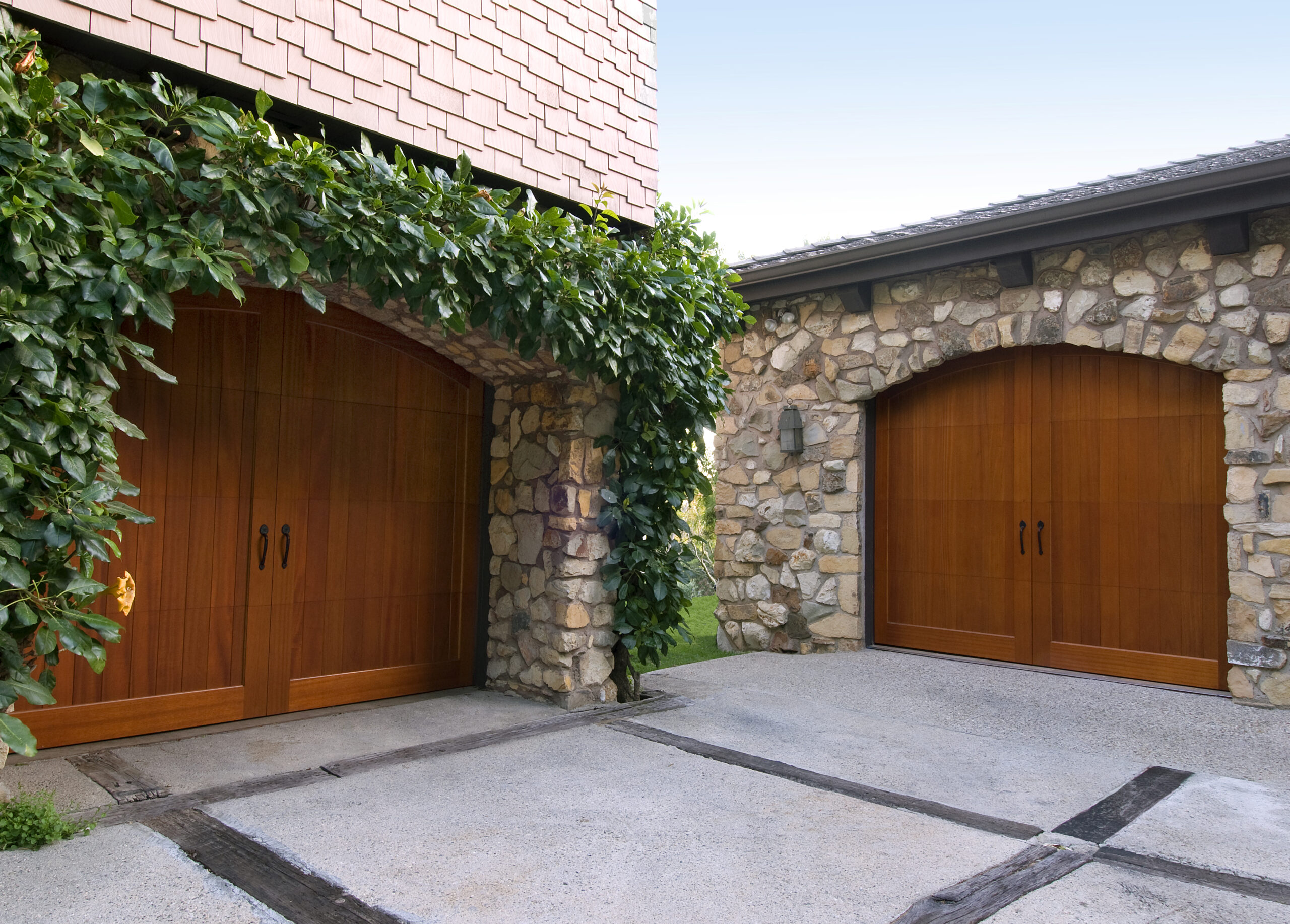 Two wooden arched garage doors with stone exteriors and greenery surrounding the entrance.