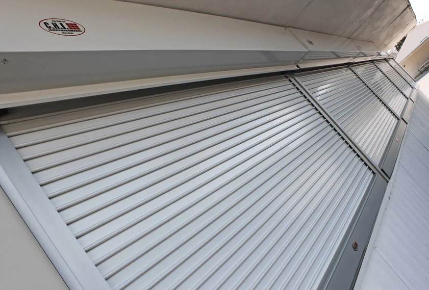 A large silver metal security roller shutter is partially open, showcasing its horizontal slats. The C.H.I. logo is visible on the housing unit above.